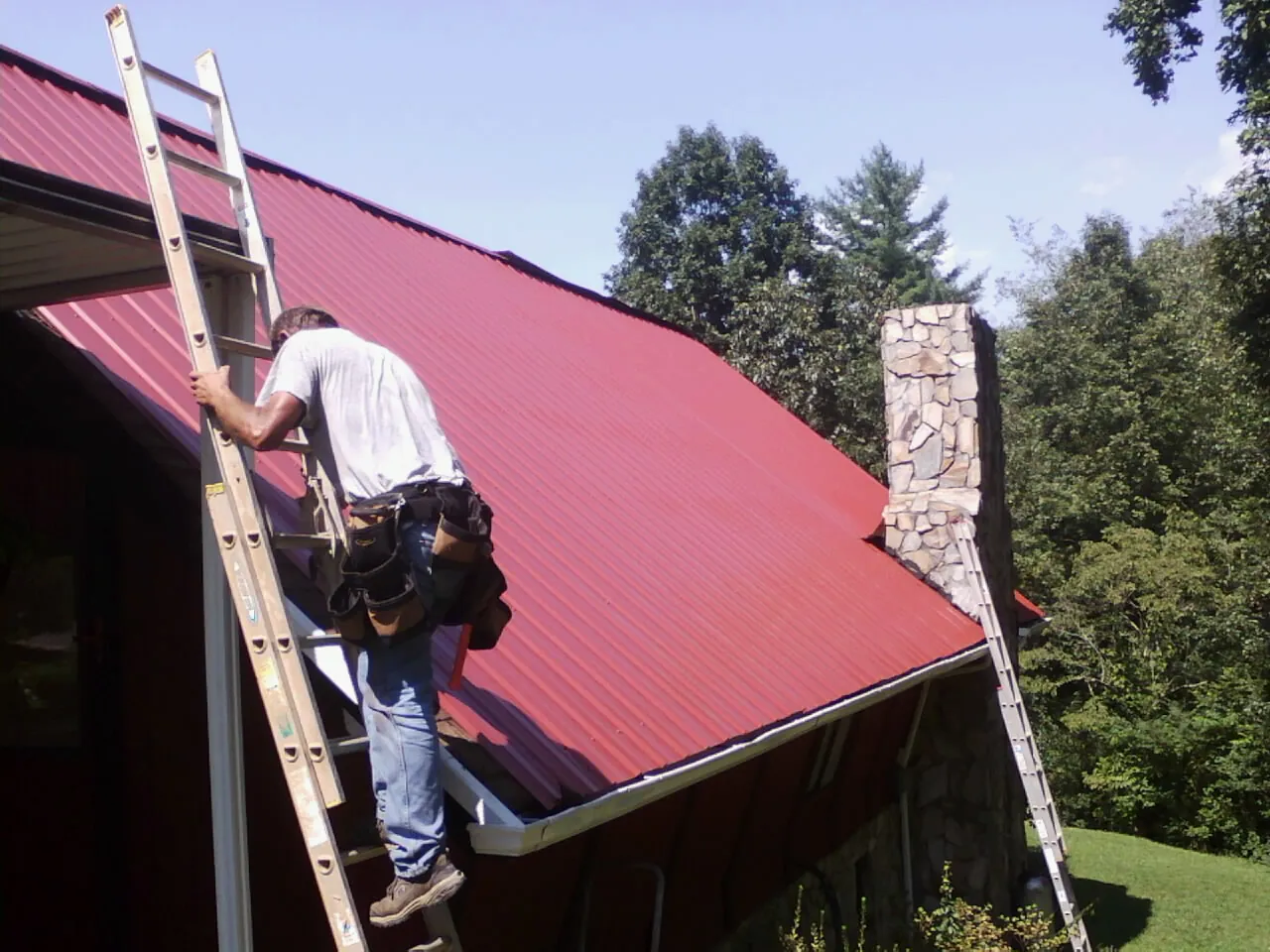 Roofer on a ladder beside a red metal roof and stone chimney — Soffit & Fascia Repair by Roofing Brothers in Ozawkie
